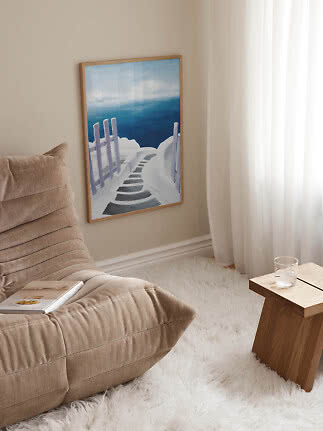 Framed painting of a white pathway by a blue sea on a beige wall beside curtains and an armchair