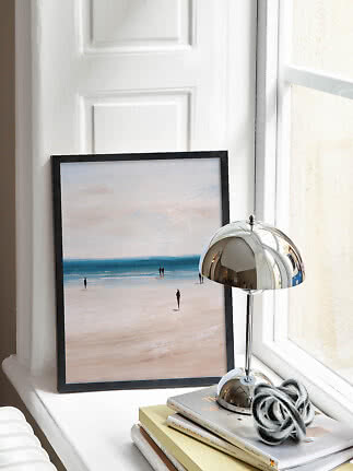 Framed beach print on a windowsill beside a chrome table lamp and stacked books
