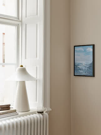 Lámpara de mesa blanca en el alféizar junto al radiador y pequeño cuadro marino en pared beige