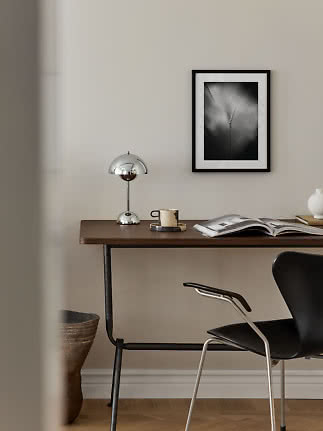 Scrivania minimalista con lampada cromata, tazza, libro aperto e sedia nera su parete beige