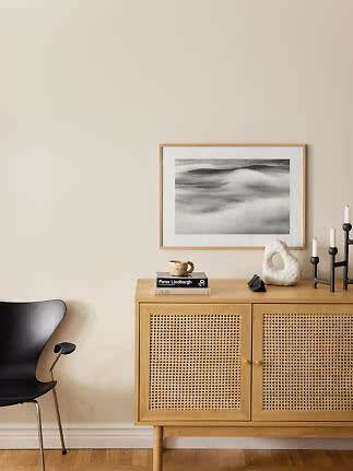 Aparador de madera clara con puertas de rejilla, silla negra y decoración bajo cuadro en blanco y negro