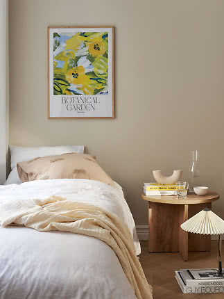 Soveværelse med indrammet botanisk plakat over sengen og rundt træbord med bøger og glasvase.
