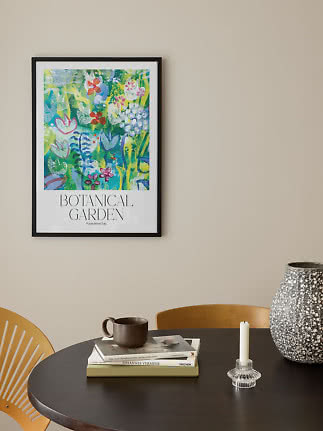 Poster botanico colorato incorniciato su parete beige sopra tavolo rotondo con vaso, candeliere, libri e tazza
