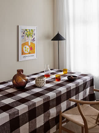 Table à manger avec nappe à carreaux bruns, vase en verre, tasses et bol dans une pièce lumineuse.