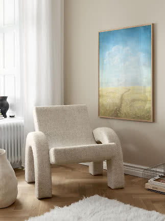 Blød beige loungestol i et lyst hjørne ved siden af et indrammet landskabsmaleri.