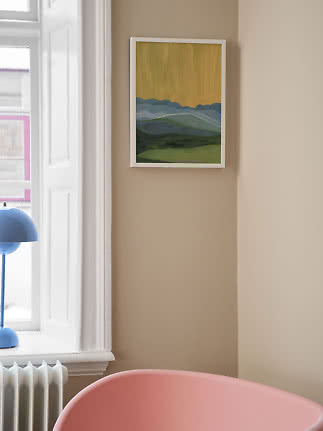 Framed landscape painting with blue mountains and green fields on a beige wall