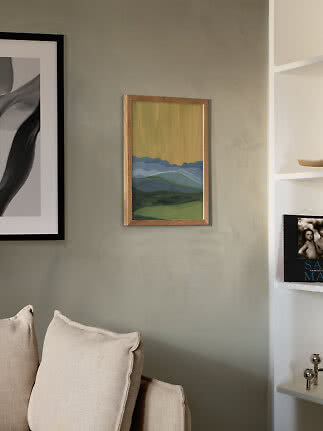 Small framed landscape painting with blue mountains and yellow sky on a green wall