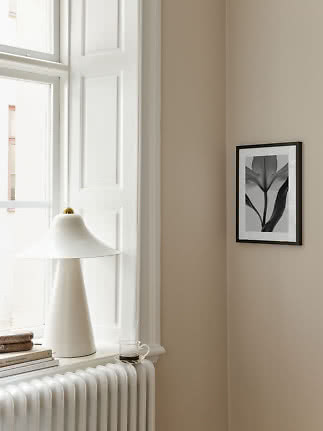Lille sort indrammet blomsterplakat på beige væg ved vindue og hvid bordlampe