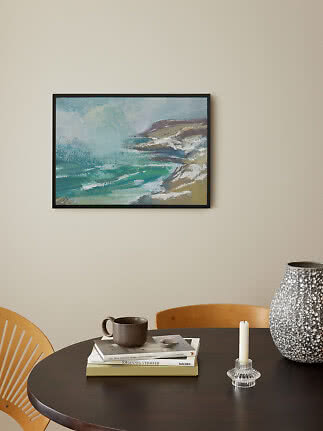 Ingelijst kustschilderij met golven aan beige muur boven ronde tafel met boeken, mok, kandelaar en vaas