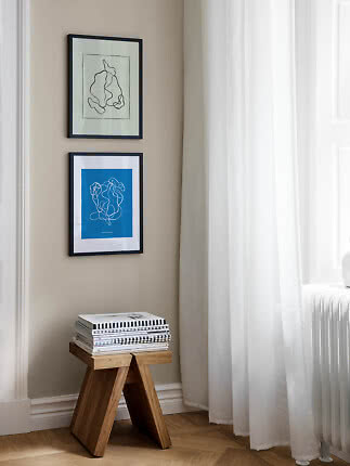 Two framed abstract prints above a wooden stool with magazines beside a window with white curtains