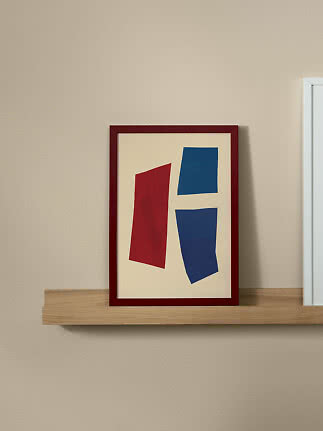 Abstrakt poster i röd ram med röda och blå former på en trähylla mot beige vägg