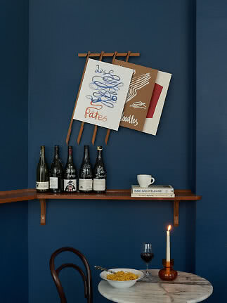 Dunkelblaue Wand mit Holzregal, Weinflaschen, Postern und kleinem Tisch mit Pasta, Weinglas und Kerze