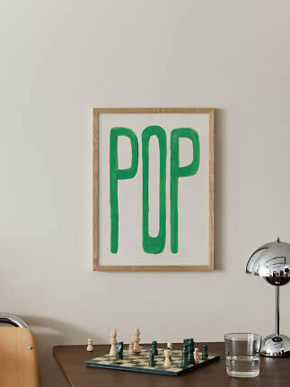 Inramad poster med grön "POP"-text på ljus vägg ovanför ett bord med schackbräde.