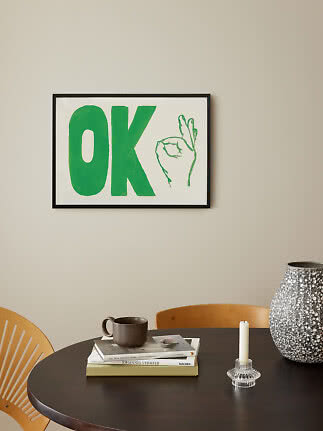 Gerahmtes Poster mit grünem „OK“ und Handzeichnung an heller Wand über rundem Tisch.