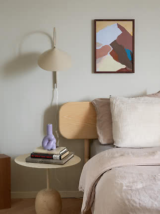 Lampada da parete beige accanto al letto con tavolino rotondo, libri, vaso viola e quadro astratto