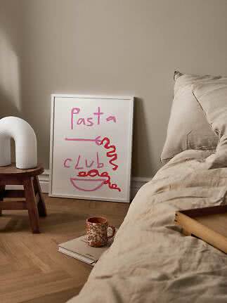 Indrammet Pasta Club-plakat lænet mod væg ved seng med beige sengetøj og lille træskammel