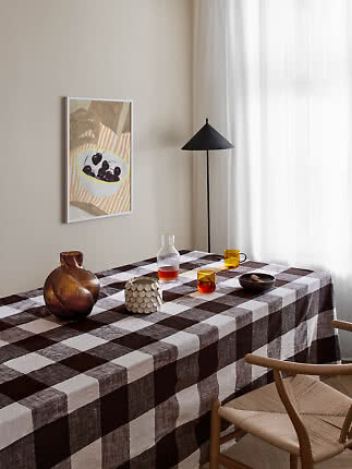 Mesa de comedor con mantel de cuadros marrón, vasos, jarrón y lámpara de pie negra junto a la ventana