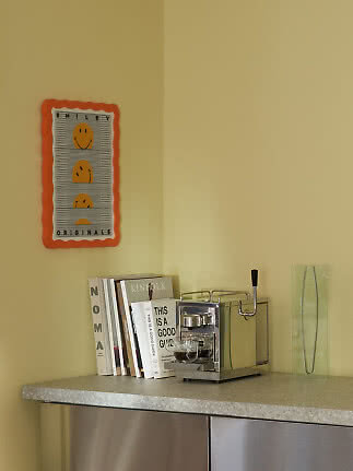 Køkkenhjørne med orange smiley-plakat, bøger, espressomaskine og høj glasvase.