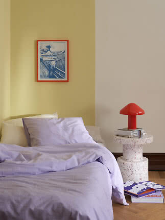 Bedroom with bed in light purple bedding, red table lamp, and framed art on a yellow wall