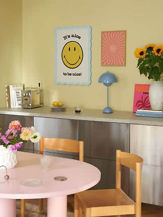 Angolo pranzo dai toni pastello con tavolo rosa, sedie in legno, lampada blu e poster smiley