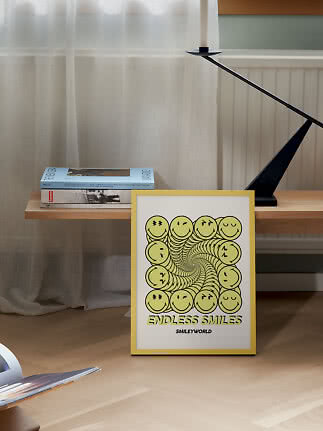 Poster SmileyWorld con cornice gialla e spirale di smiley, appoggiato a una panca in legno con libri e portacandela nero