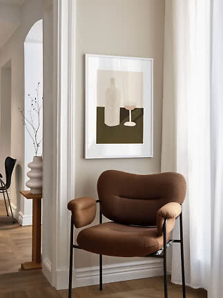 Brun polstret stol ved beige væg med indrammet stillebenplakat og hvid gardin