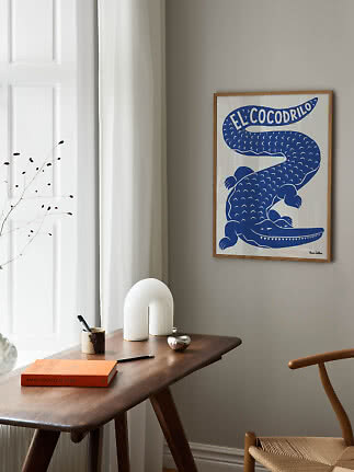 Gerahmtes Poster mit blauem Krokodil an beiger Wand über einem Holztisch am Fenster
