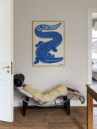 Blaues Krokodil-Poster mit „El Cocodrilo“ in hellem Holzrahmen über einem Loungesessel.