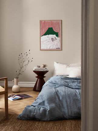 Soveværelse med lyseblåt sengetæppe, lille sidebord, vase og farverigt indrammet kunsttryk på væggen