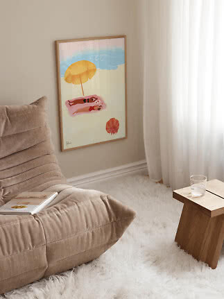 Framed beach poster with two people on a towel under a yellow umbrella in a bright room corner