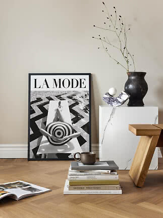 Stylish interior with framed La Mode poster, vase with branches, marble pedestal and stack of books.