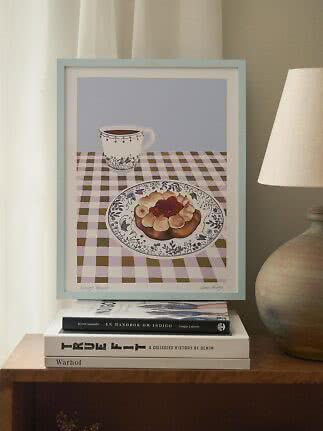 Stampa da cucina incorniciata con tazza e dolce su tovaglia a quadri, su libri accanto a una lampada