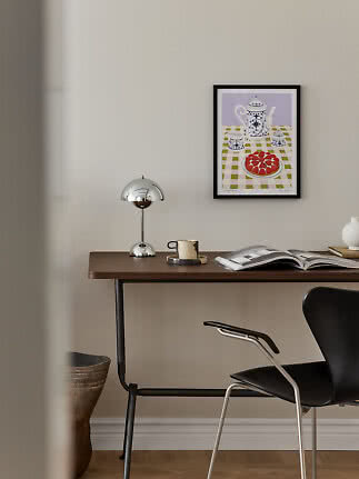 Minimalistisch bureau met chromen lamp, mok, open boek en ingelijste keukenprint aan de muur