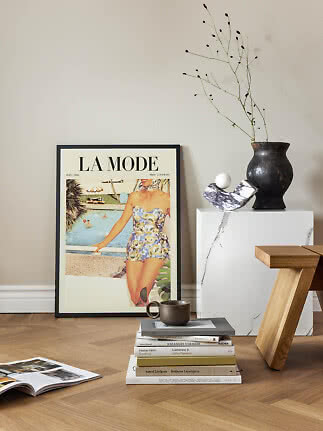 Framed vintage fashion poster leaning against wall with books, cup, black vase and wooden stool