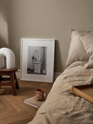 Hyggeligt soveværelse med beige sengetøj, hvid lampe på skammel og indrammet kunst på gulvet