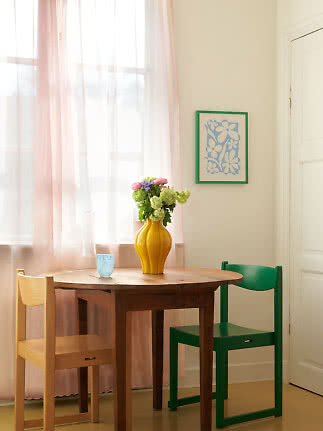 Dining nook with round wooden table, yellow flower vase and two chairs by pink curtains