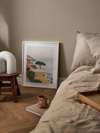 Cozy bedroom with beige bedding, framed coastal print and white lamp on a wooden stool