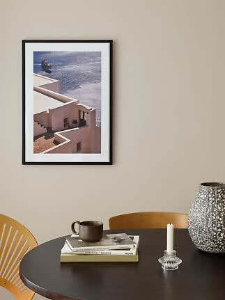 Black-framed coastal wall print above a dark table with vase, candle holder, books and mug