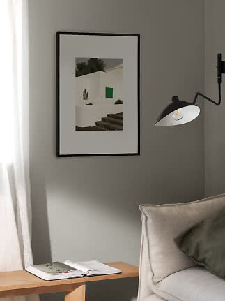 Black framed print of a white building with green window on a gray wall beside sofa and lamp