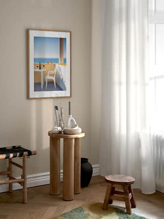Framed seaside poster above a round wooden table by a sunlit window