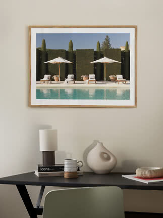 Indrammet plakat med pool, liggestole og hvide parasoller på en beige væg