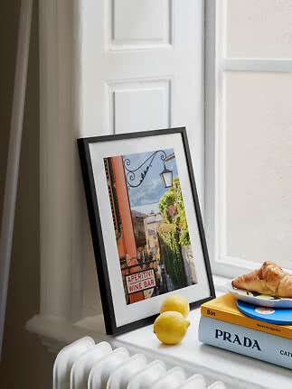 Lámina en marco negro junto a una ventana con limones, libros y un cruasán
