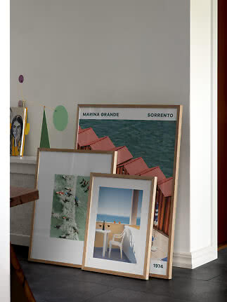 Three framed art prints with light wood frames leaning against a gray wall in a modern home.