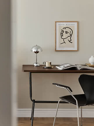 Skrivebord med kromlampe, kop, åben bog og sort stol under indrammet Matisse-plakat.
