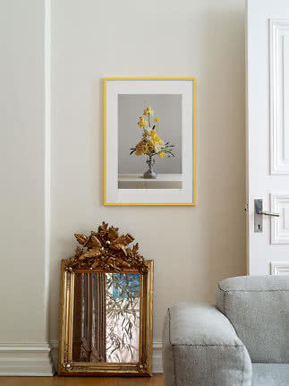 Gele lijst met bloemenposter aan beige muur boven een sierlijke gouden spiegel.