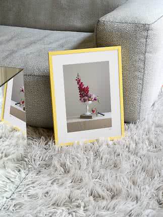 Goudkleurige fotolijst met bloemenprint, leunend tegen een grijze bank op wit vloerkleed