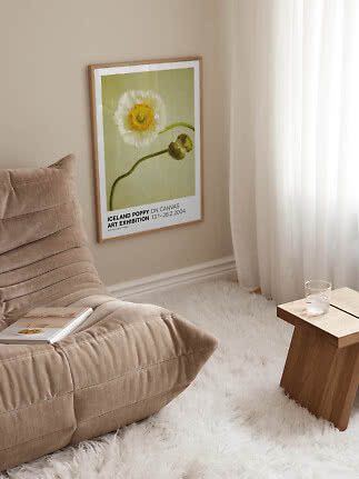 Framed floral poster with white poppy on beige wall beside lounge chair and small wooden table