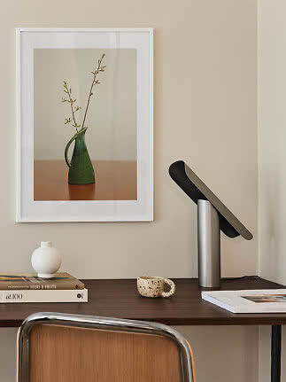 Modern desk with silver table lamp, books, mug and framed print of a green vase on a beige wall