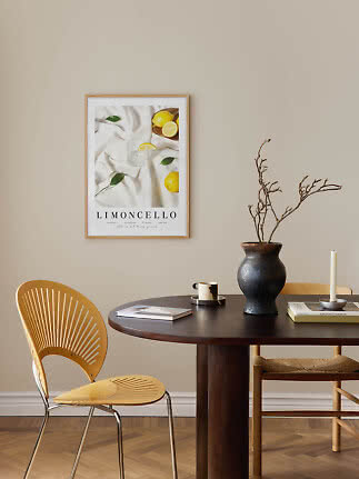 Limoncello-poster in houten lijst boven donkere eettafel met vaas, kandelaar en stoelen