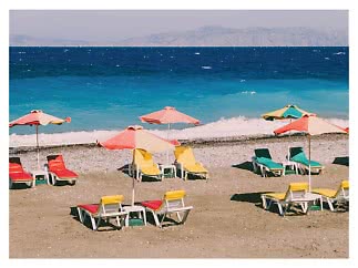 Bunte Sonnenschirme und Liegen am Strand vor türkisblauem Meer mit Bergen im Hintergrund
